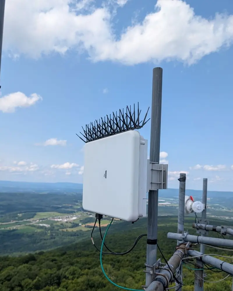 A modern base-station radio mounted on a tower mast against a blue sky, with a valley and ridges below.