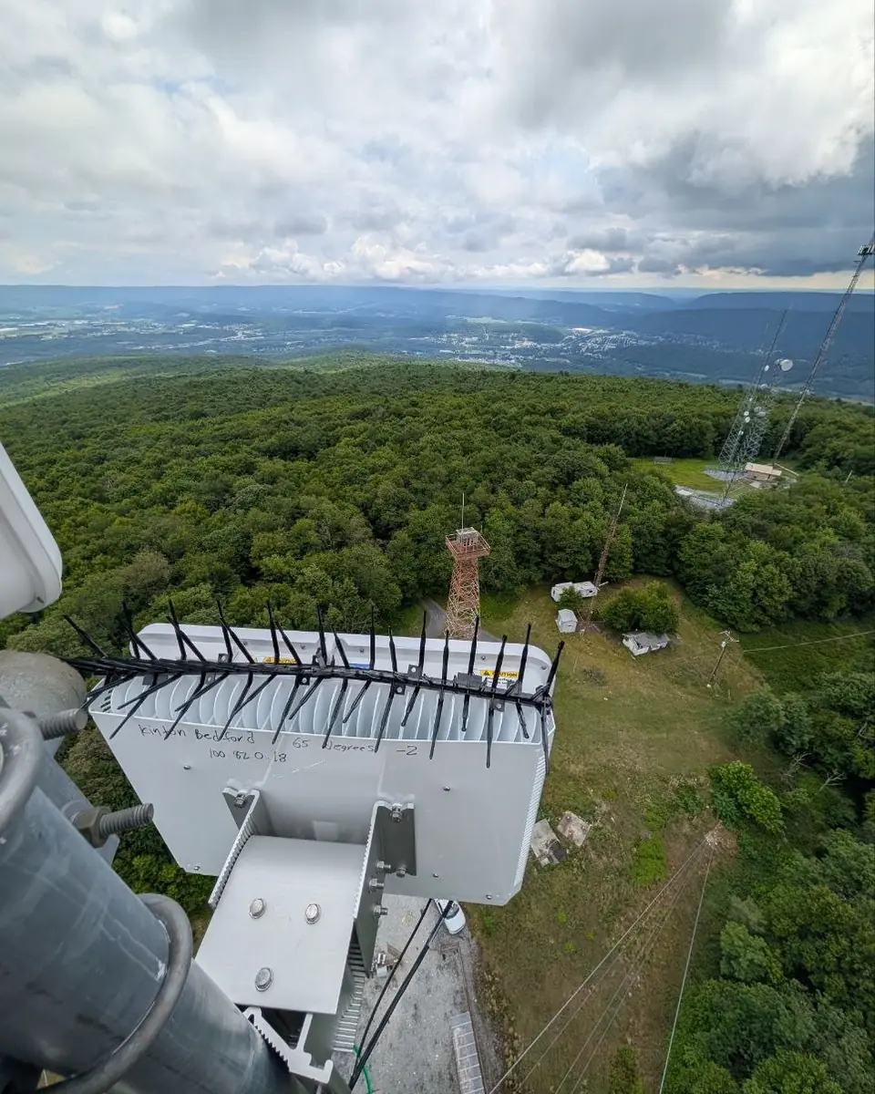 A modern base-station radio on a tower above a forested summit and a wide valley under dramatic clouds.