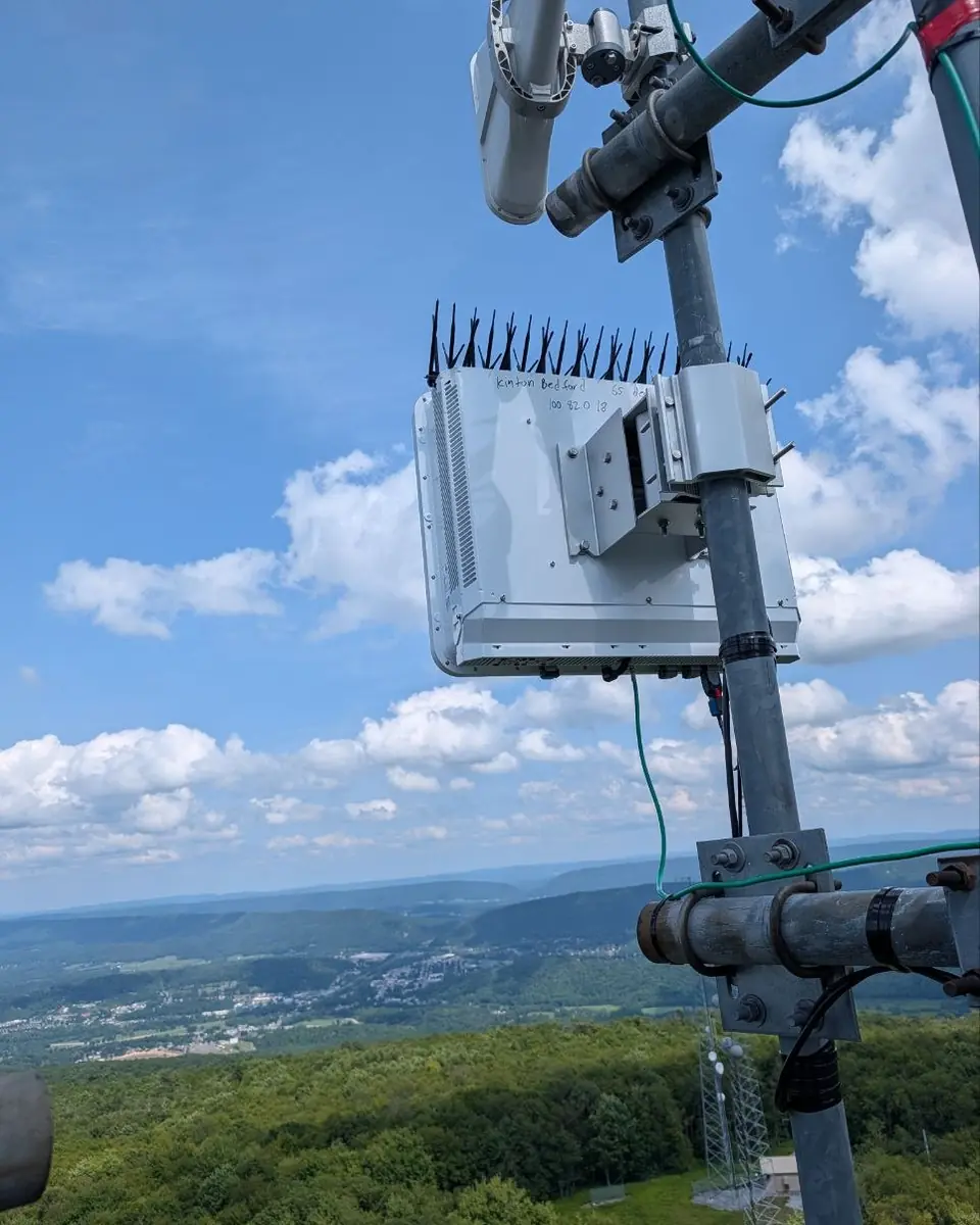 A modern base-station radio on a tower mast against a blue sky with clouds, a valley town below.