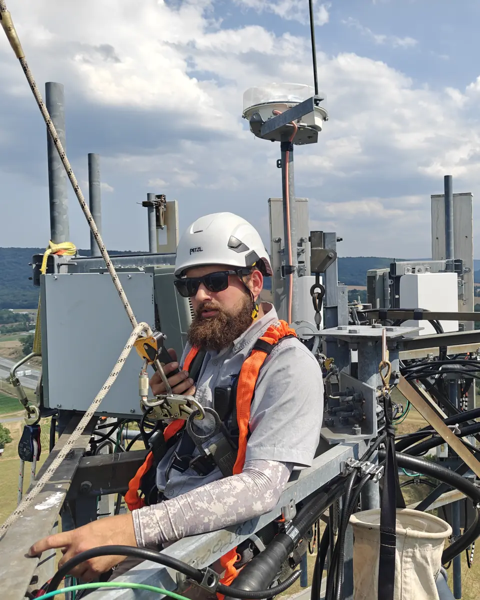 A Crowsnest tower technician in a helmet and harness on a tower platform among radios, with farmland and ridges far below.