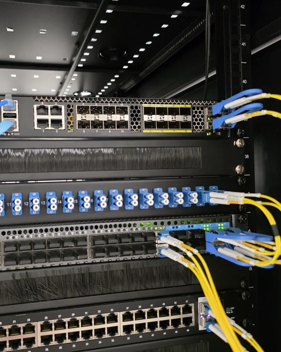 Inside a Crowsnest core network rack: routers, fiber patch panels, and yellow fiber jumpers under rack lighting.