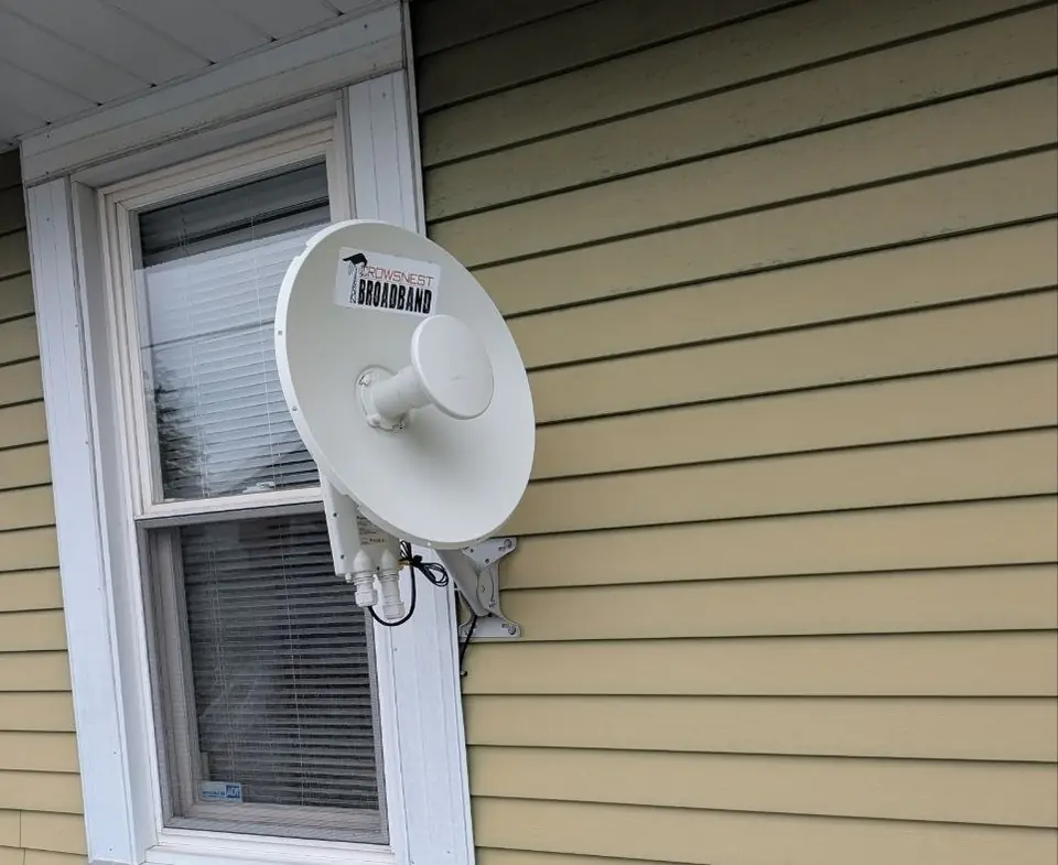 A round customer antenna on a clean wall mount beside a window on yellow siding.