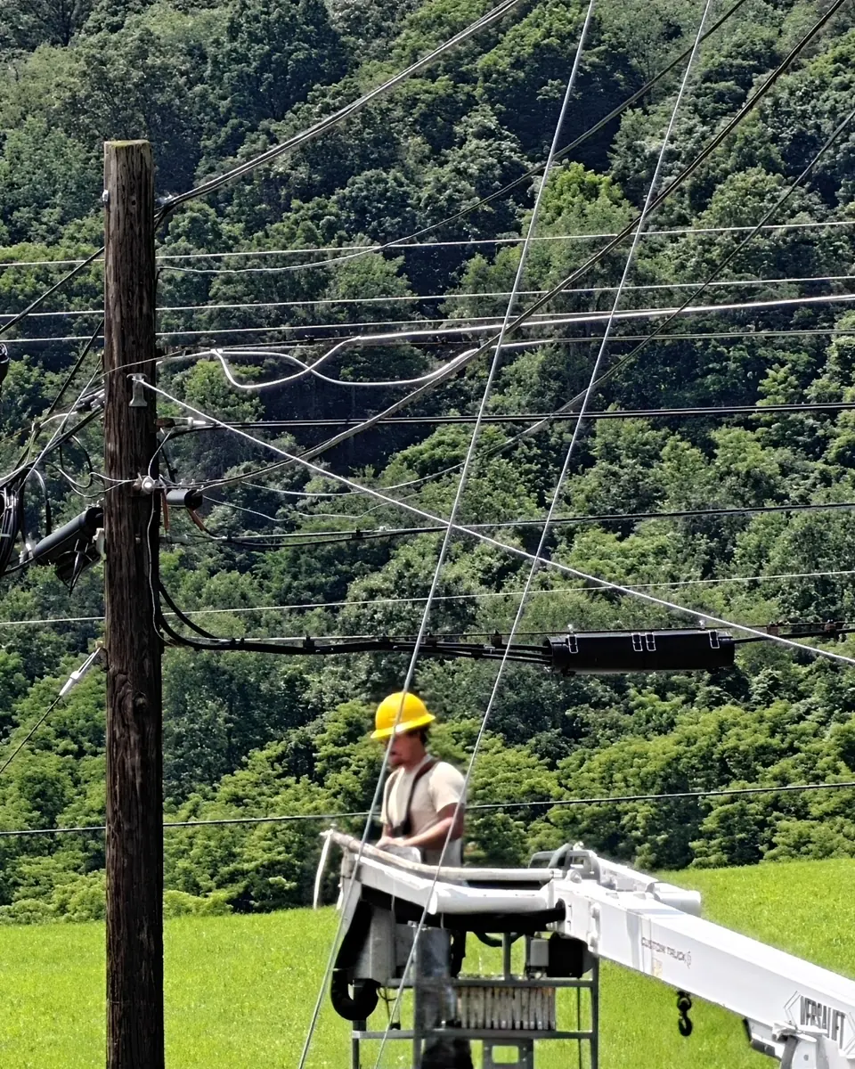 A Crowsnest lineman in a hard hat working from a bucket beside a utility pole with fiber closures, green forest behind.