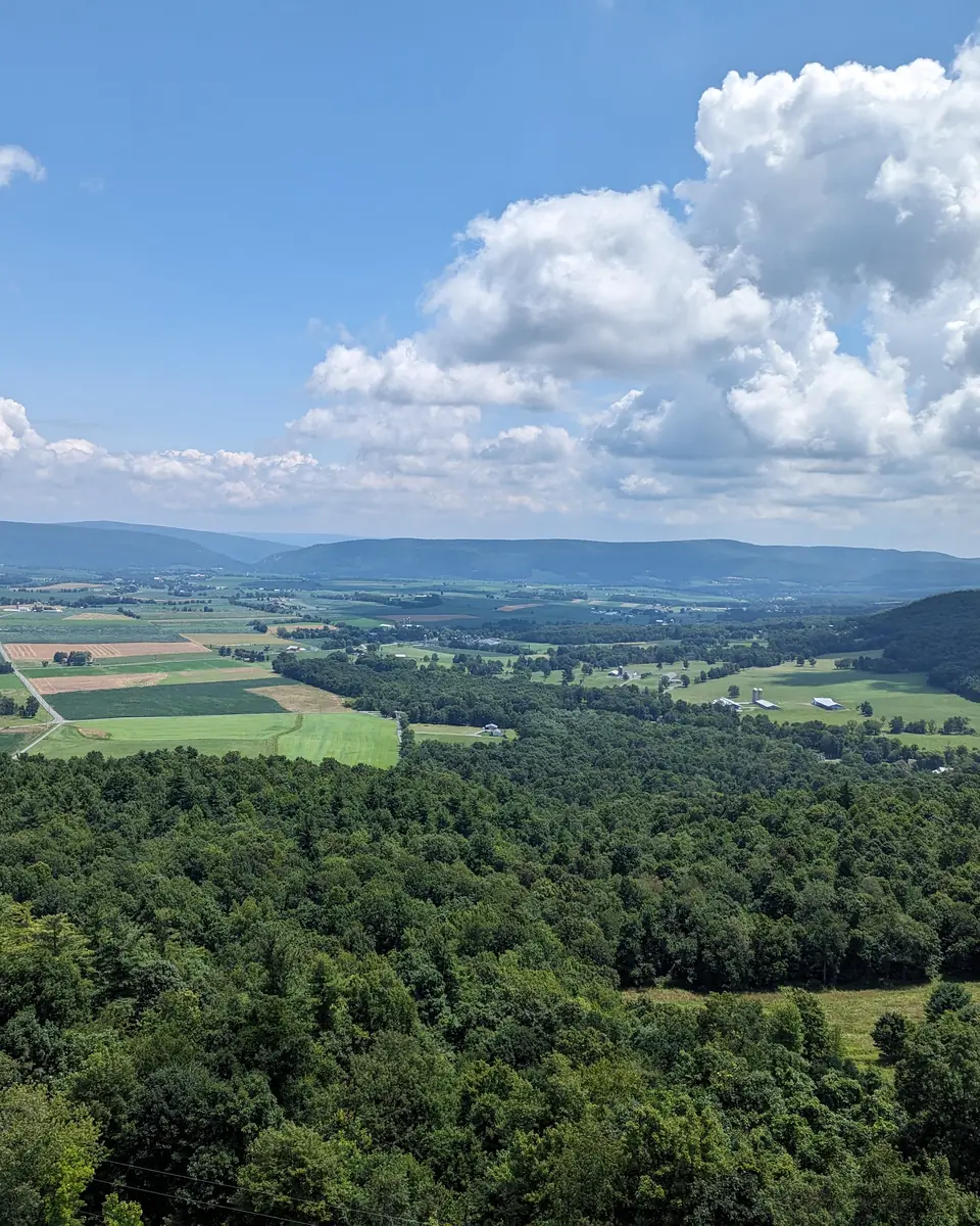 A bright summer valley of Central Pennsylvania farm fields and wooded ridges under big white clouds.