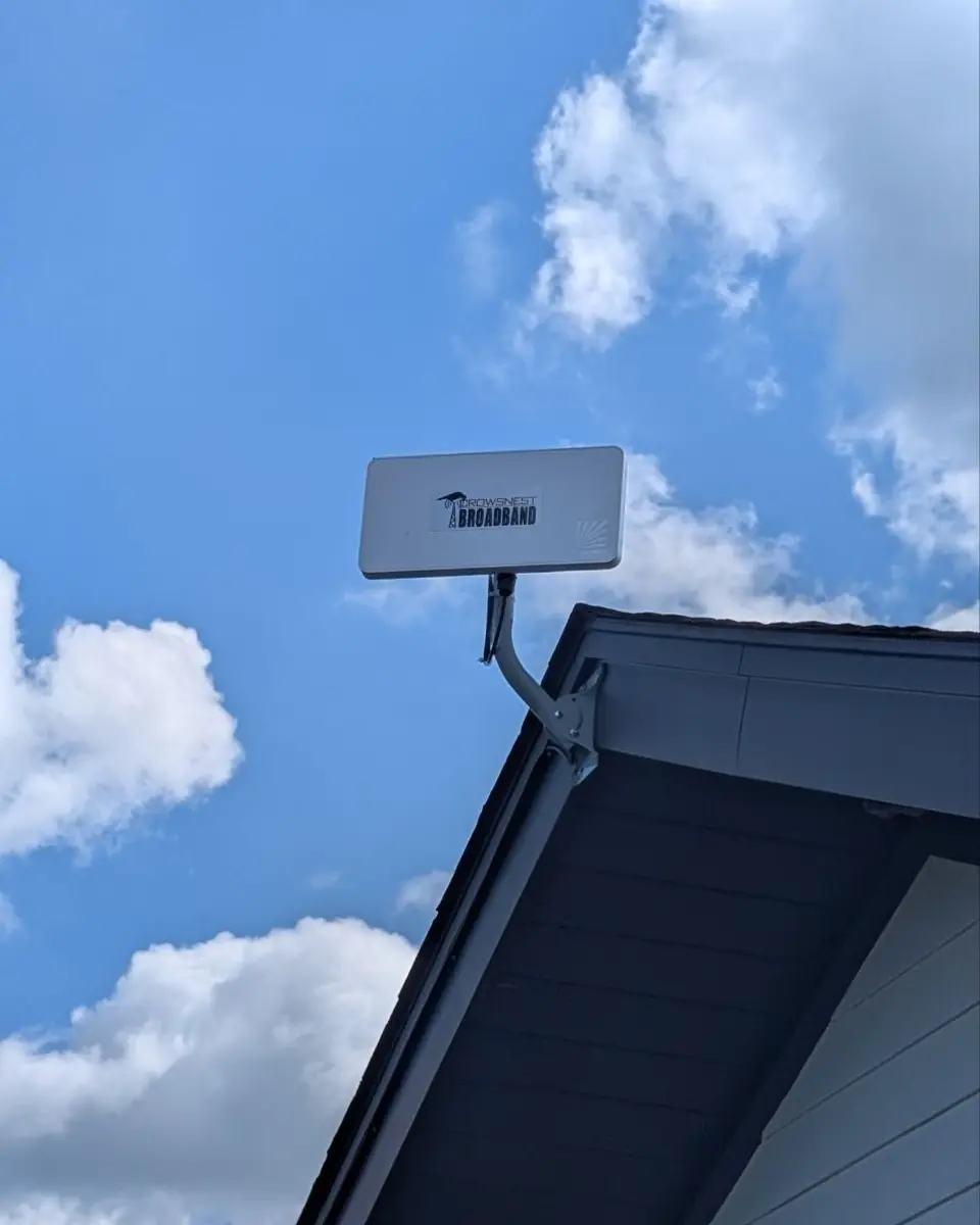 A Crowsnest-branded Tarana radio neatly mounted on a gable bracket against a blue sky with white clouds.