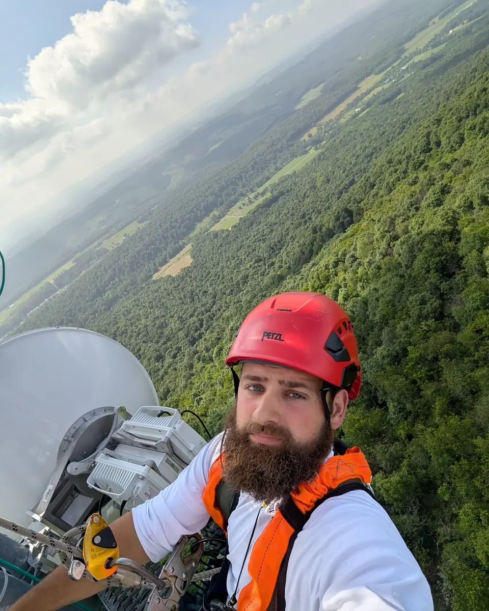 A bearded Crowsnest technician in a red helmet and harness high on a tower above a forested ridge and valley.